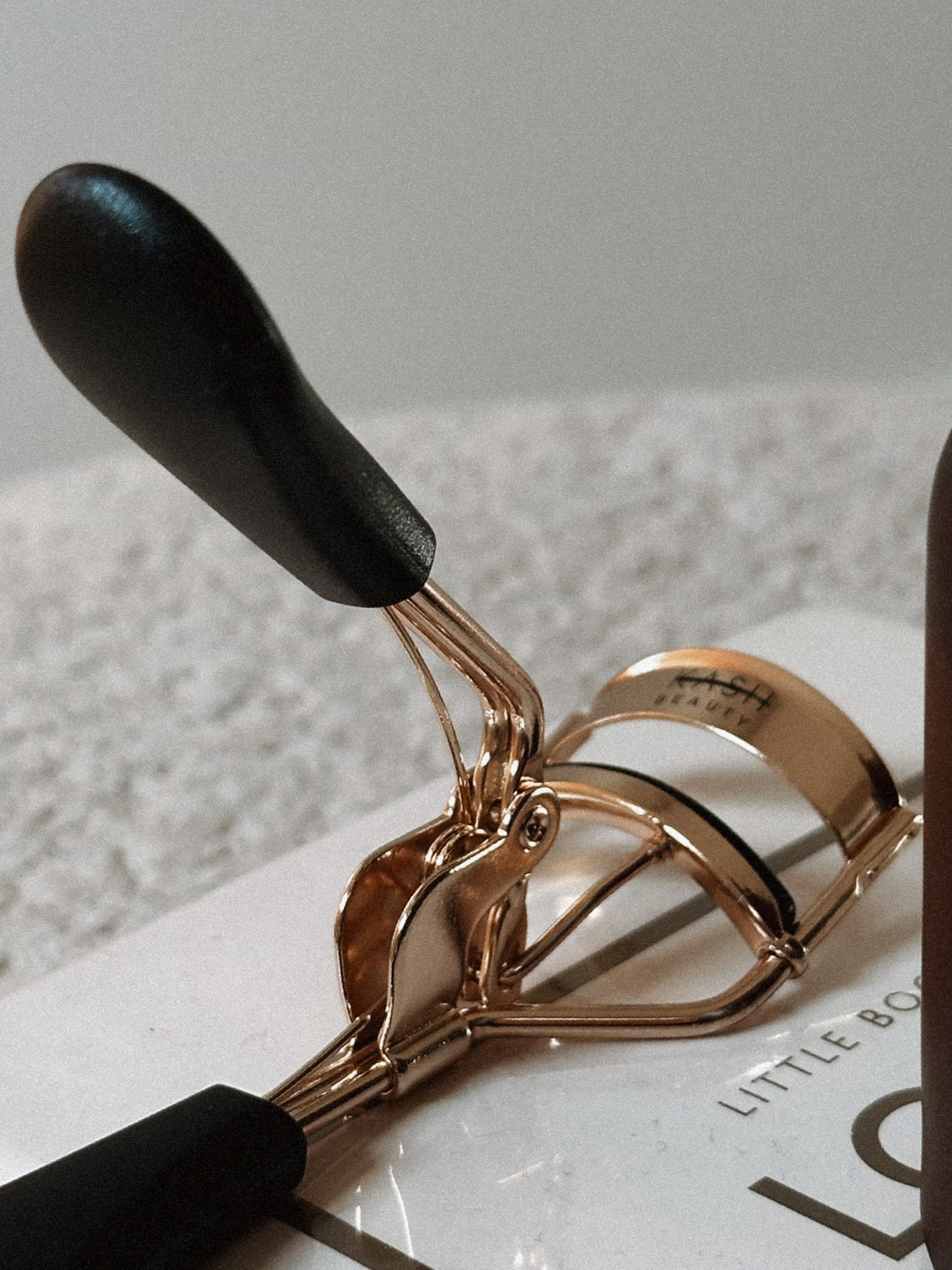 Gold eyelash curler with black handles on a light textured surface, beauty tool close-up.