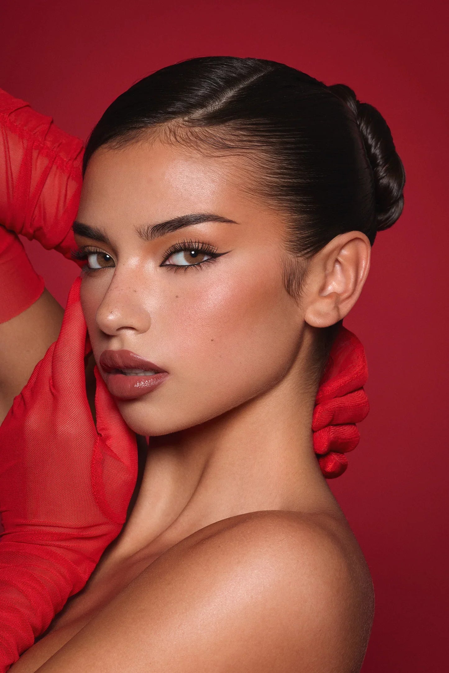 Woman with flawless makeup, winged eyeliner, and glossy lips, posing in red gloves against a red background