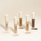 Five liquid concealers in various skin tones with swatches on a beige background