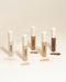 Five liquid concealers in various skin tones with swatches on a beige background