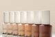 Row of Aura Skin Radiant Foundation bottles in multiple shades on a neutral background