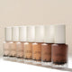 Row of Aura Skin Radiant Foundation bottles in multiple shades on a neutral background
