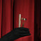 Black gloved hand holding a gold eyelash curler and mascara against red curtain