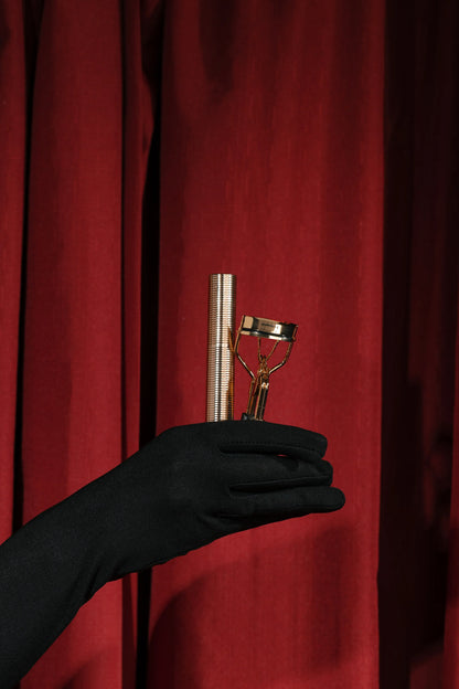 Black gloved hand holding a gold eyelash curler and mascara against red curtain