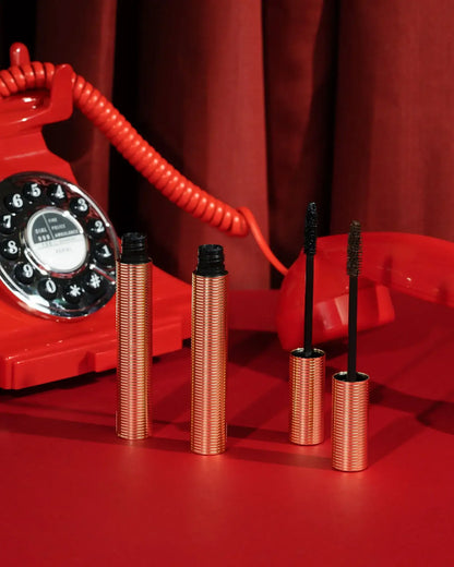 Two metallic gold mascara tubes with brushes open on a red background beside a vintage red phone.