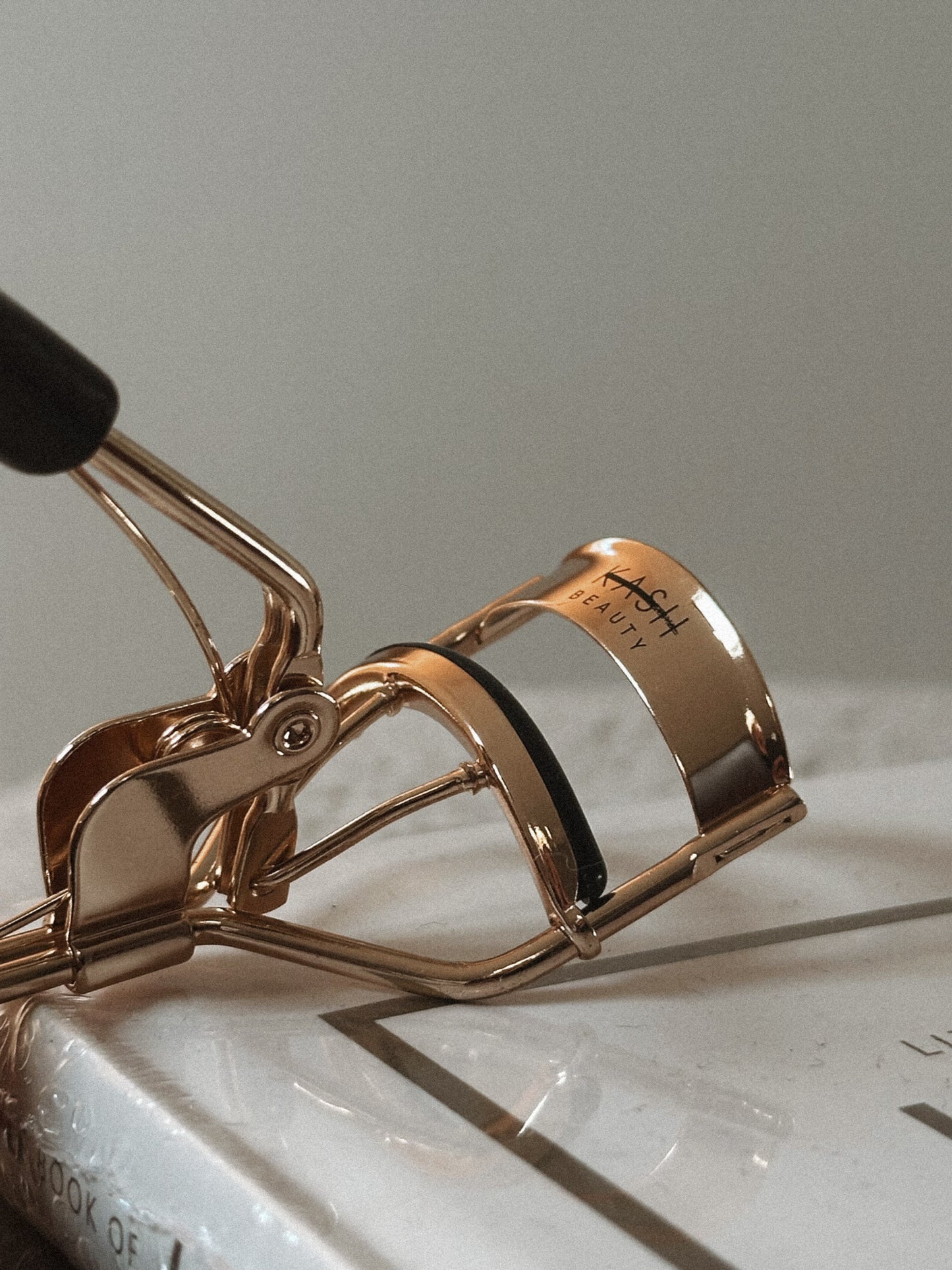 Gold metal eyelash curler with black handle on white book, beauty tool close-up