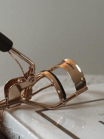Gold metal eyelash curler with black handle on white book, beauty tool close-up