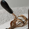 Gold eyelash curler with black handles on a light textured surface, beauty tool close-up.