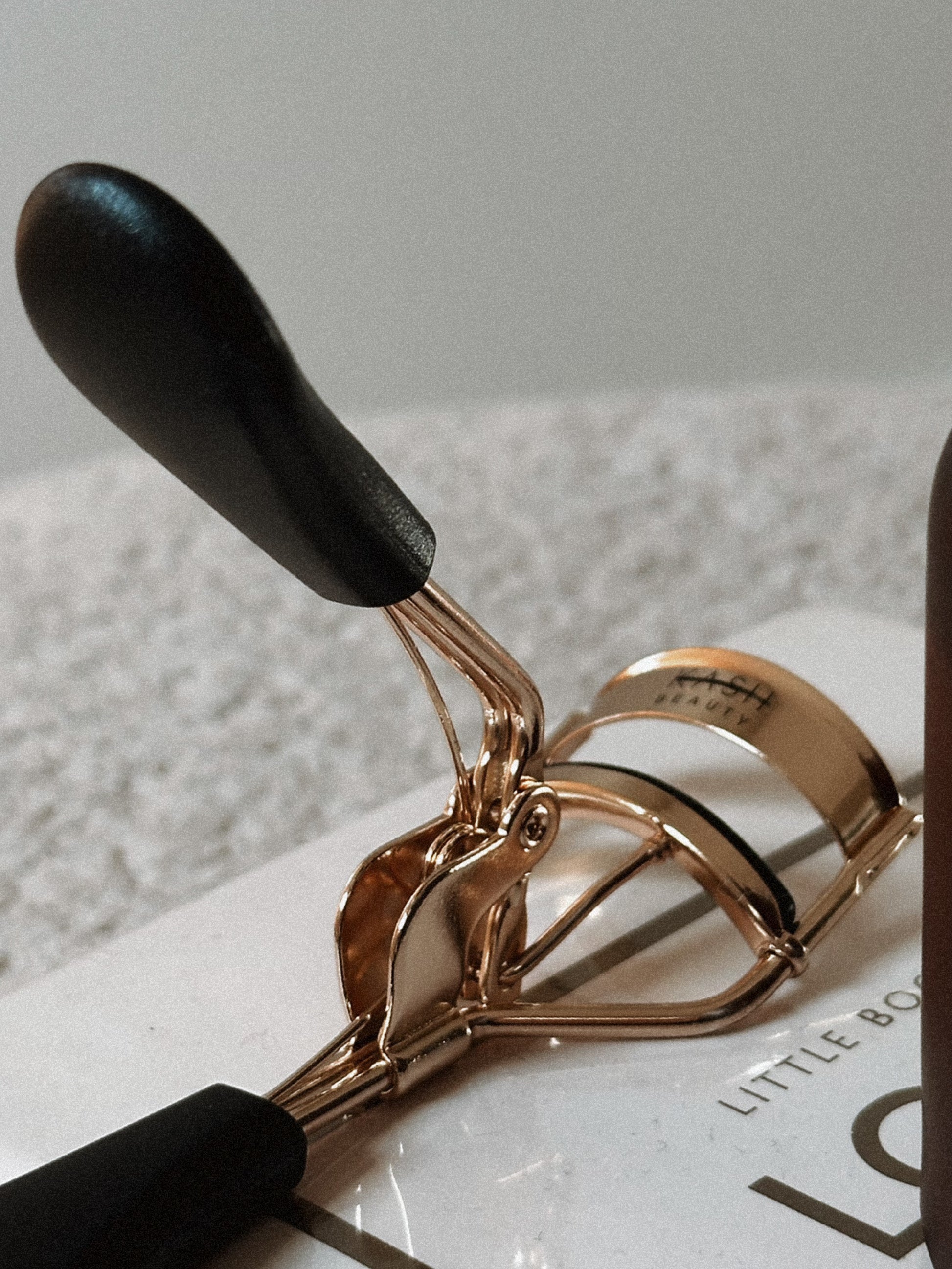 Gold eyelash curler with black handles on a light textured surface, beauty tool close-up.