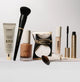 Makeup set with primer, foundation, powder, concealer, mascara, and brushes on white background.