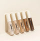 Five liquid concealers in different shades lined up on a beige background