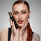 Woman with red braided hair and tattoos holding black and gold makeup bottle, flawless glam look