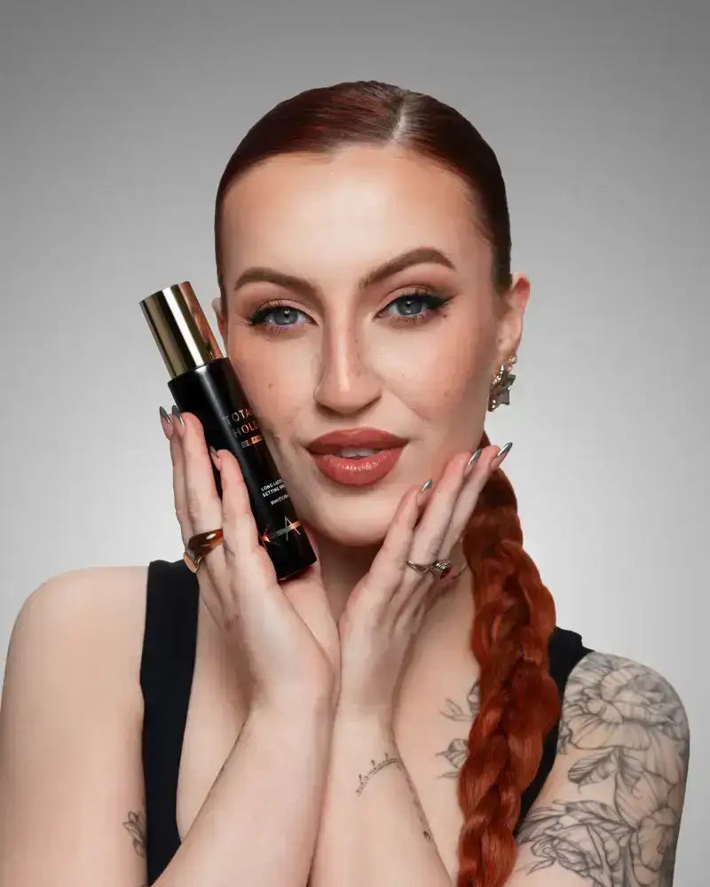 Woman with red braided hair and tattoos holding black and gold makeup bottle, flawless glam look